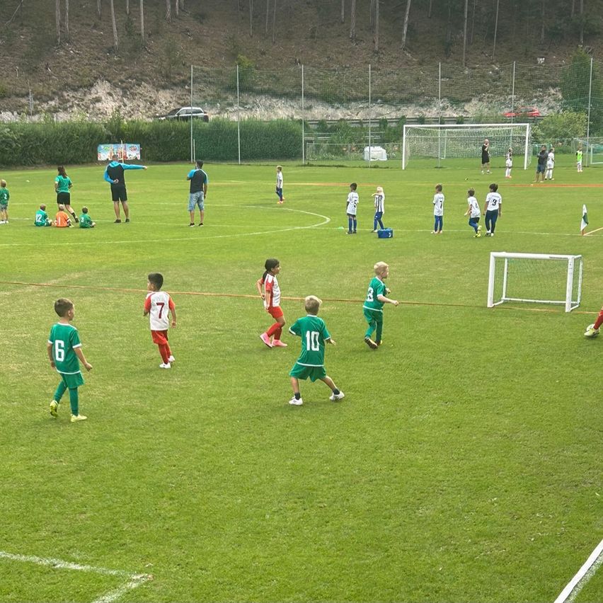 Kinder in grünen und weißen Uniformen spielen Fußball auf einem Feld mit Toren an jedem Ende. Einige rennen, während andere stehen und zuschauen.