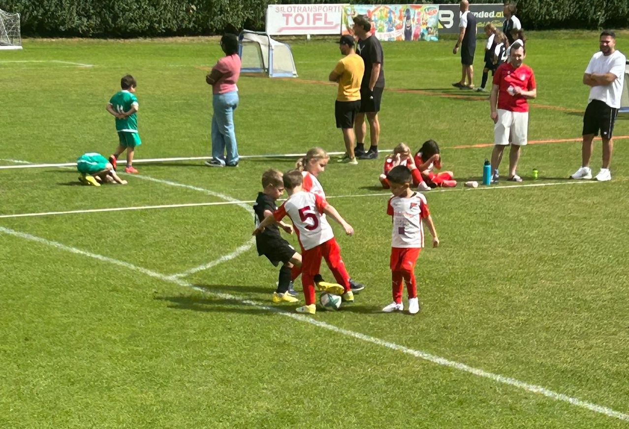 Eine Gruppe von Kindern spielt Fußball auf einem Feld, während Erwachsene zuschauen. Ein Junge in einem roten Trikot mit der Nummer 5 läuft auf den Ball zu.