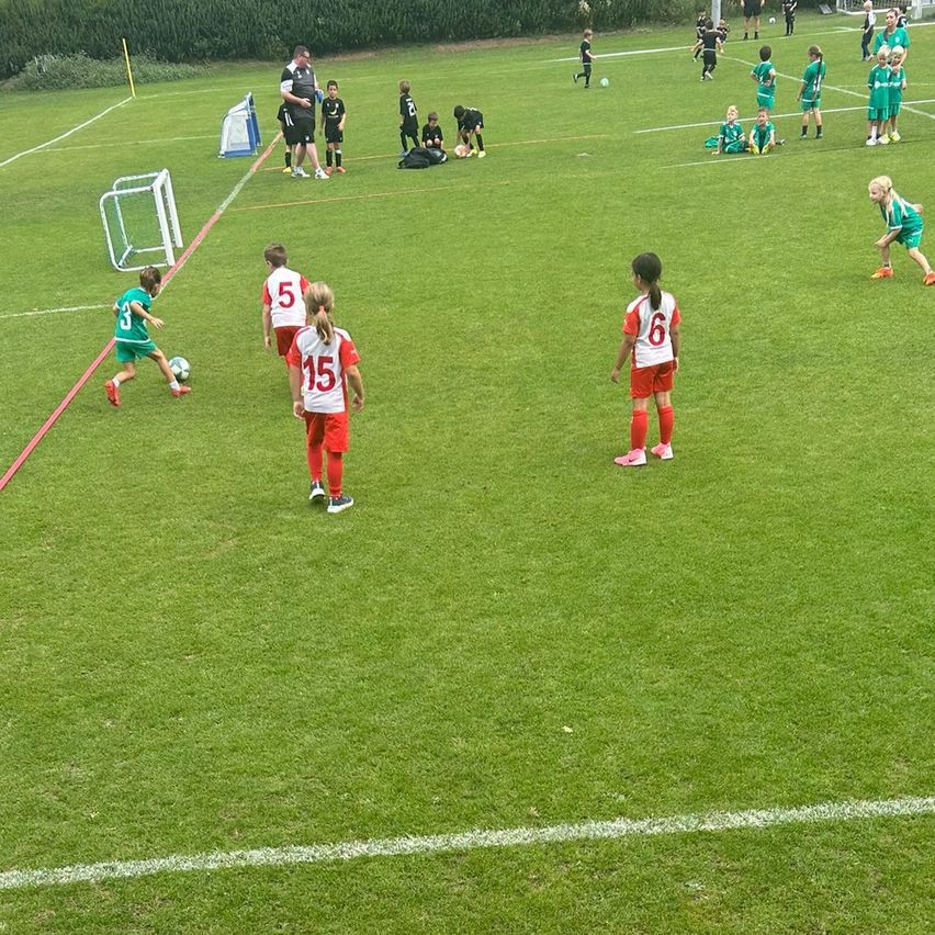 Eine Gruppe von Kindern in grünen und roten Uniformen spielt Fußball auf einem Feld. Einige stehen, während andere sitzen oder knien. Das Feld hat weiße Markierungen und ein Tor.