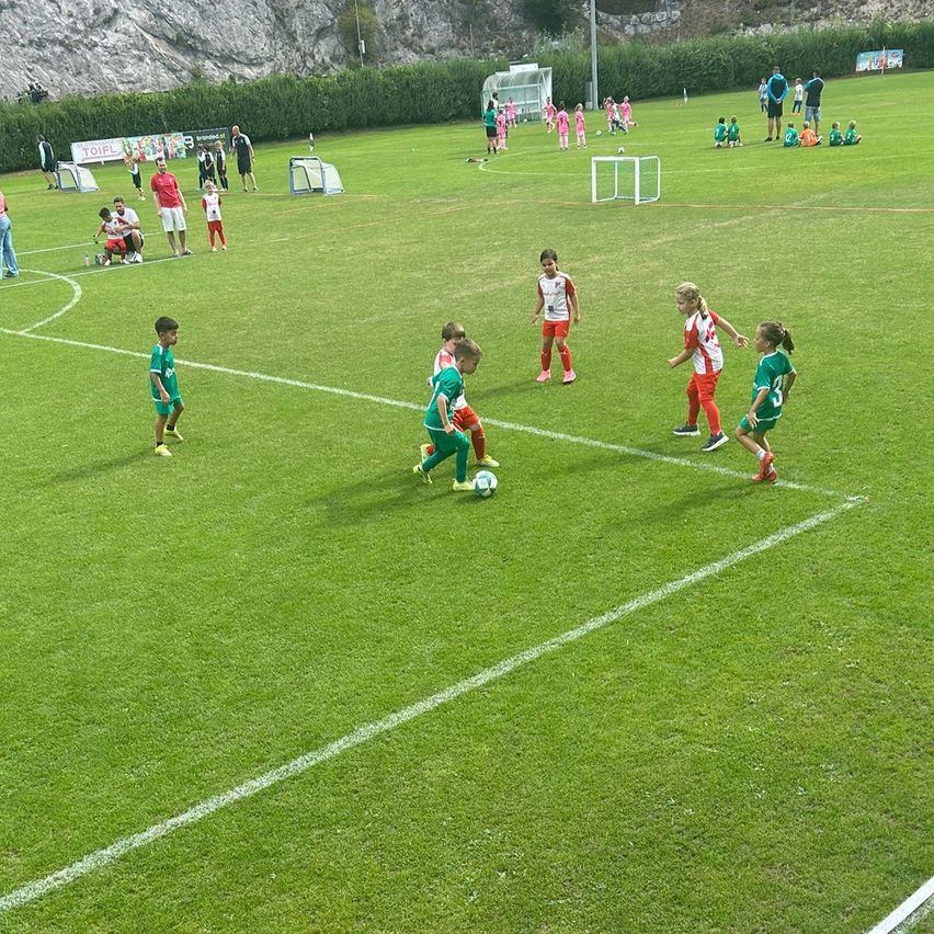 Eine Gruppe von Kindern in grünen und roten Trikots spielt Fußball auf einem Feld mit weißen Markierungen. Einige laufen auf den Ball zu, während andere in der Nähe der Torpfosten stehen. Im Hintergrund befinden sich weitere Kinder und Erwachsene, einige stehend, einige sitzend.