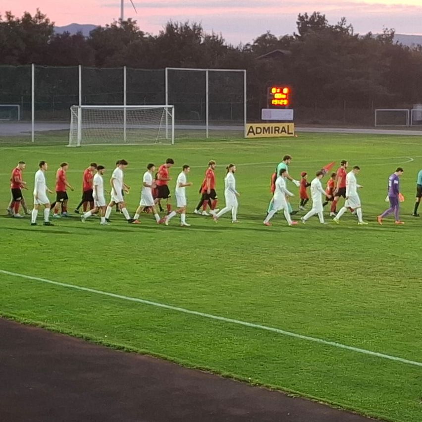 A soccer team in white uniforms is walking off the field at dusk, with a scoreboard and sponsor sign visible in the background.