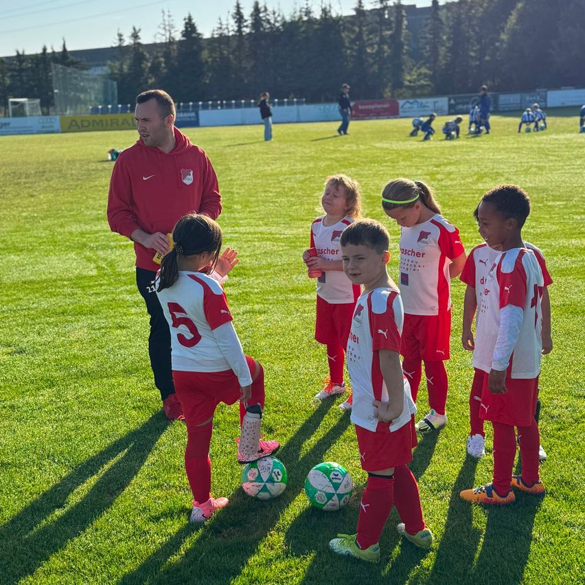 A soccer coach in a red hoodie instructs a group of young players on a green field. The children, wearing red and white uniforms, are attentively listening.