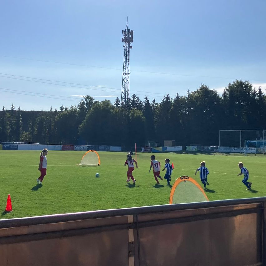 A group of children playing soccer on a field with a goal net, a tower in the background, and a clear blue sky.
