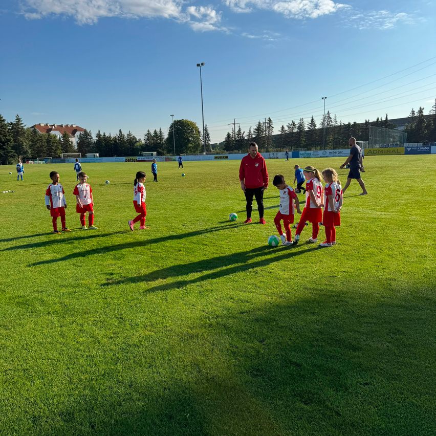 A group of children in red and white uniforms are practicing soccer on a field with a coach in red nearby. The field is surrounded by trees and a building.