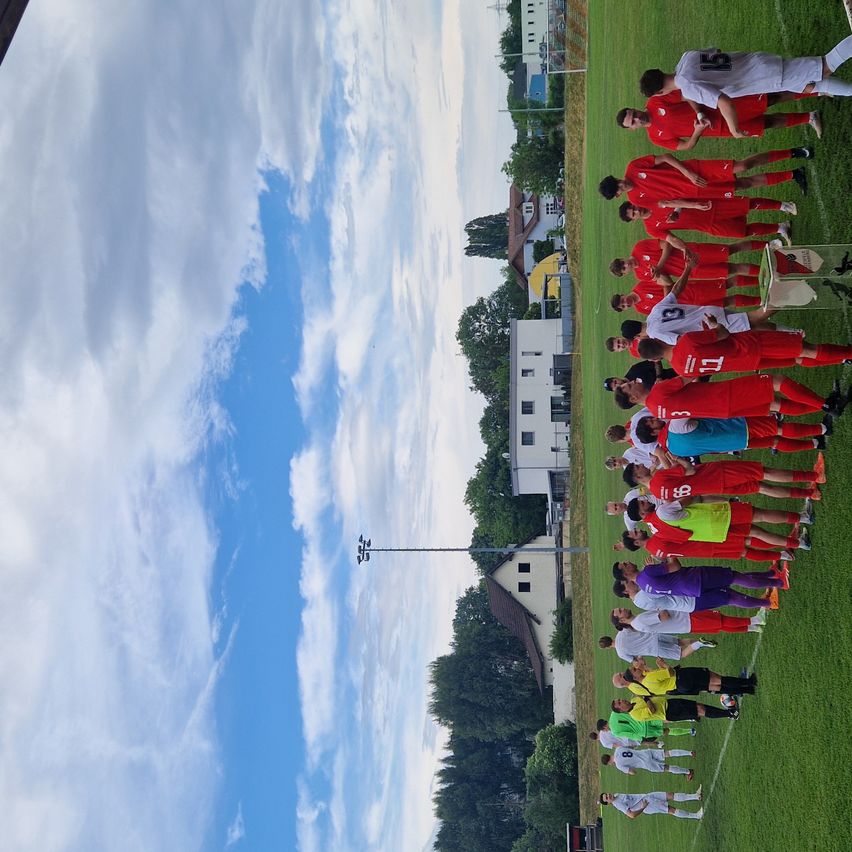 Eine Fußballmannschaft versammelt sich auf einem Rasenfeld und trägt rote Trikots. Hinter ihnen steht ein Gebäude mit einem gelben Dach, Bäumen und einem klaren Himmel mit Wolken.