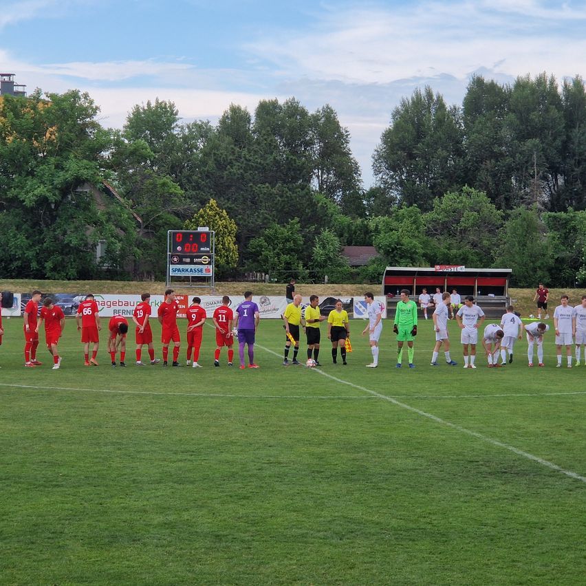 Fußballspieler in roten und grünen Trikots stehen auf einem Feld in einer Reihe. Eine Anzeigetafel zeigt 0:0. Bäume und ein Gebäude sind im Hintergrund.