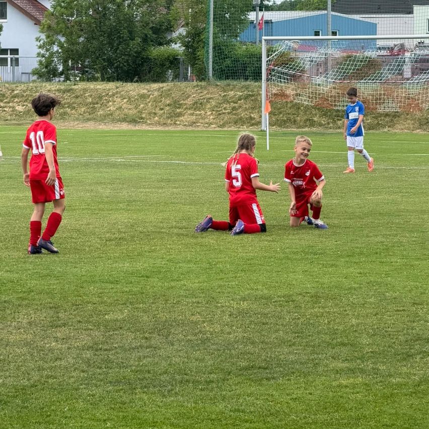 Vier Jungen in roten Trikots spielen Fußball auf einem Feld mit einem Tor. Zwei Jungen knien, während die anderen beiden stehen. Ein Junge in blauem Trikot läuft hinter dem Tor.