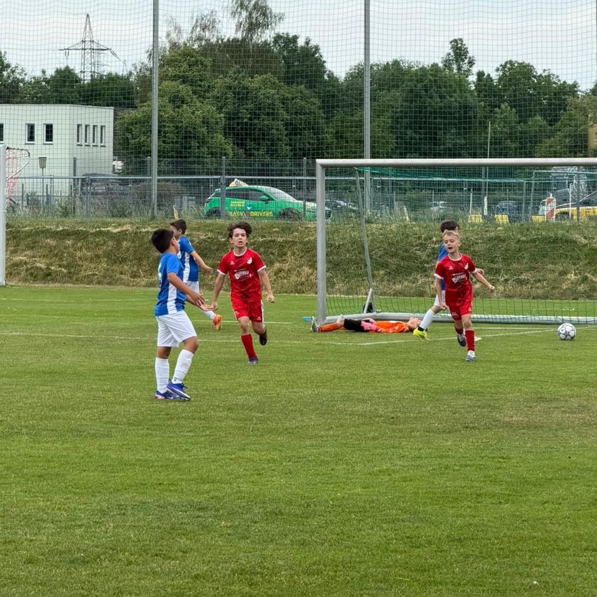 Ein Fußballspiel findet auf einem Feld statt, die Spieler tragen rote und blaue Uniformen. Der Torwart steht in der Nähe des Tores. Ein Ball liegt auf dem Feld.