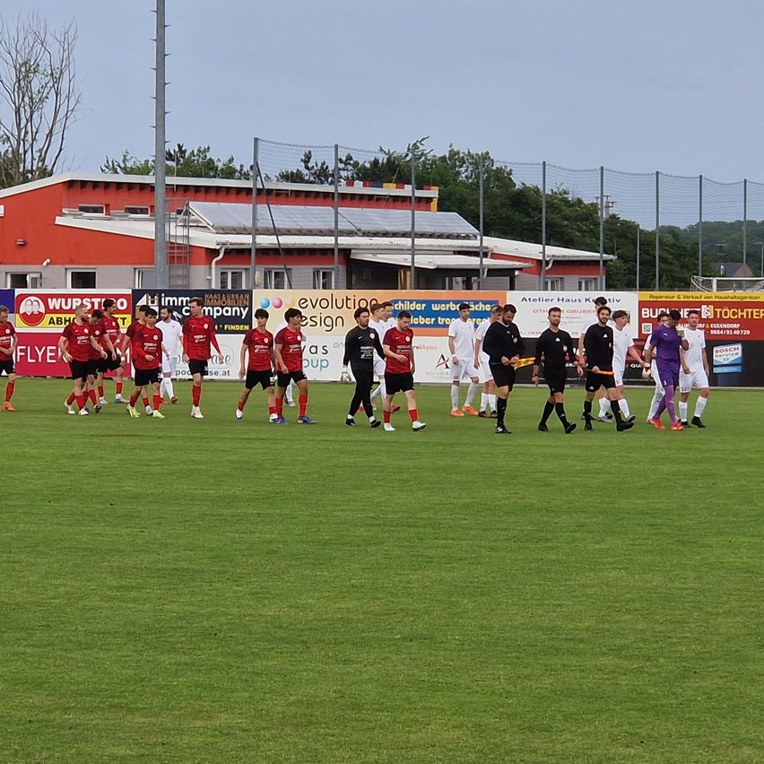 A soccer match is in progress on a field. Players in red and black uniforms walk towards the center. A red building with solar panels is in the background.