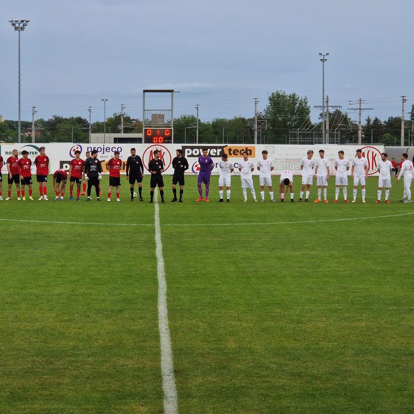 Two soccer teams are standing on the field, wearing red and white uniforms. The field has white lines. There are two referees. Behind them, there is a wall with advertisements.