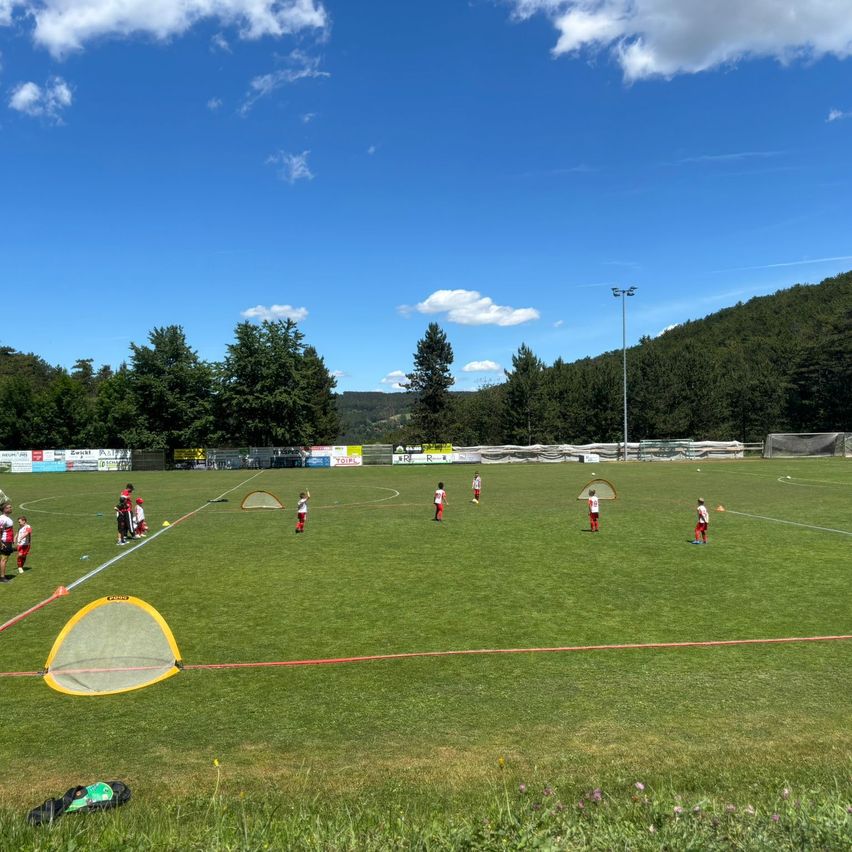 A soccer field with green grass and players in red jerseys practicing on a sunny day. A net is on the left, and several people are near the trees.