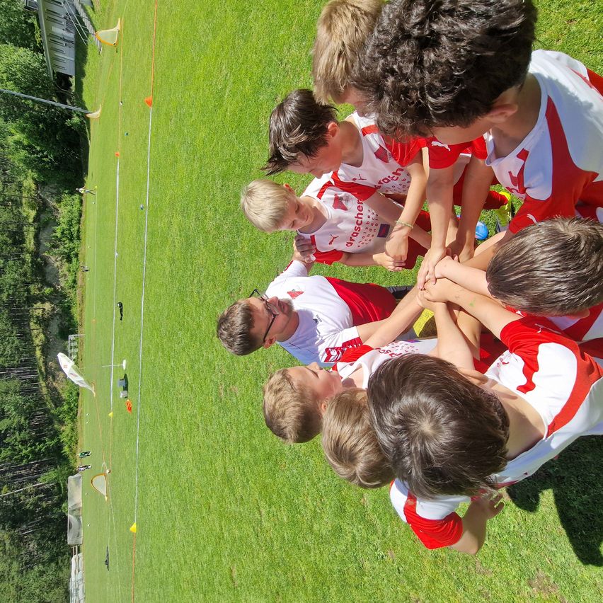 A group of children in white and red jerseys gather on a field. They are preparing for a game, with their hands clasped together. Behind them is a grassy field with orange cones.