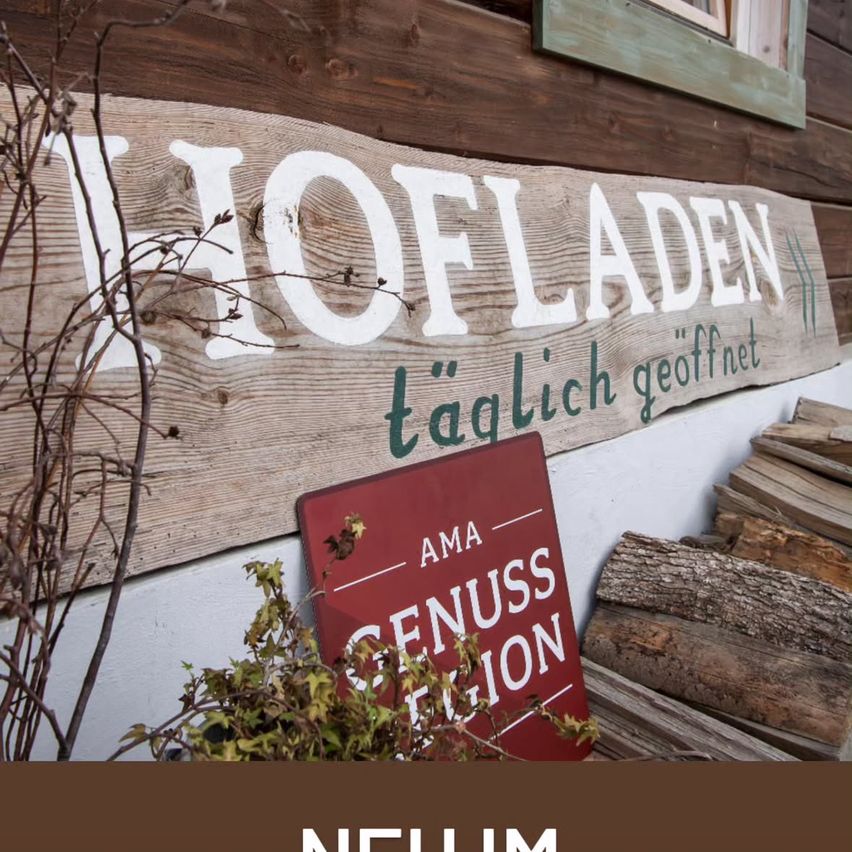 Ein Holzschild an einem rustikalen Gebäude zeigt 'HOFLADEN taglich geoffnet'. Darunter befindet sich ein rotes Schild mit weißen Buchstaben, das 'AMA GENUSS FUSION' anzeigt. Daneben liegt ein Stapel Holzscheite.