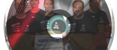 Fünf Männer stehen vor einem roten Hintergrund, alle tragen schwarze T-Shirts. Die zentrale Figur hält ein Schild mit dem Wort 'Seiwald' und einer Website-Adresse.