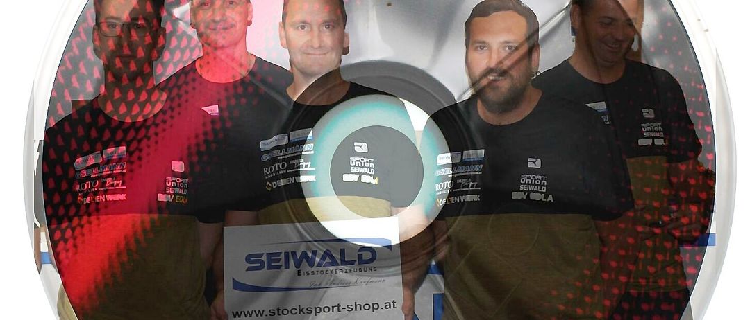 Fünf Männer in passenden T-Shirts posieren für ein Bild vor einem roten Hintergrund. Die zentrale Figur hält ein weißes Schild mit Text und einer Website. Das Bild hat einen kreisförmigen, glänzenden Abschluss.