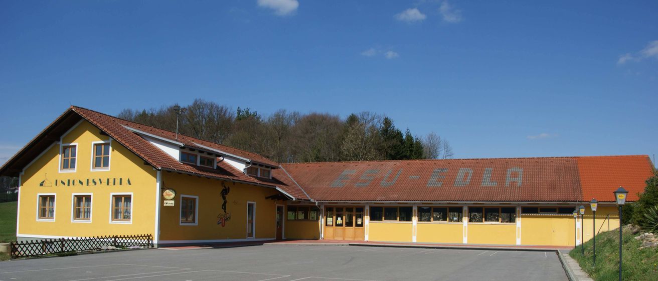 Außenansicht eines Gebäudes mit rotem Ziegeldach und gelben Wänden. Auf dem Dach steht 'ESU-ED'. Bäume sind im Hintergrund zu sehen.