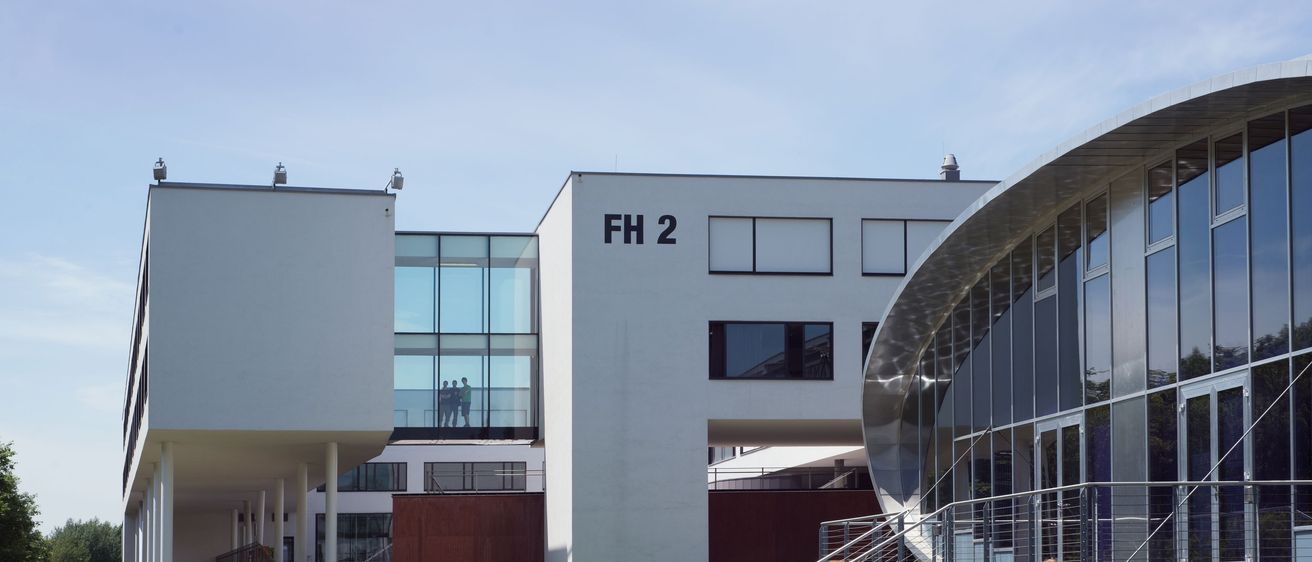 Mehrere Personen sitzen auf Bänken vor einem modernen Gebäude, das mit FH 2 beschriftet ist. Das Gebäude hat Glaswände und moderne Architektur. Ein Mann geht auf einem Kiesweg.