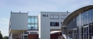 Mehrere Personen sitzen auf Bänken vor einem modernen Gebäude, das mit FH 2 beschriftet ist. Das Gebäude hat Glaswände und moderne Architektur. Ein Mann geht auf einem Kiesweg.