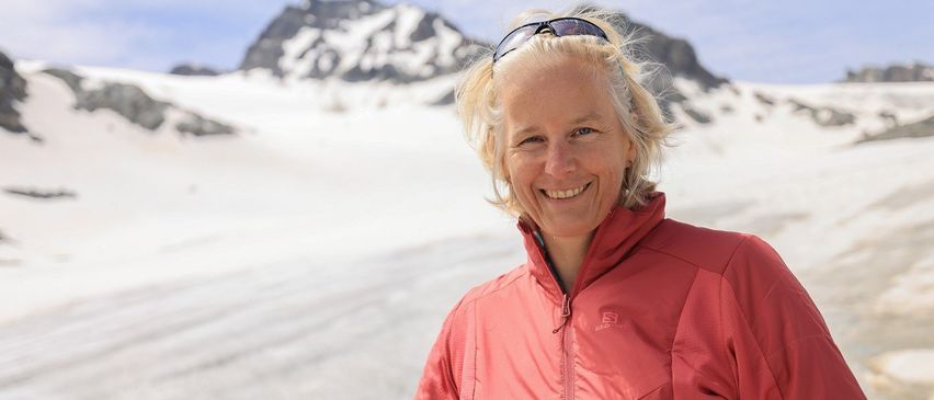 Eine Frau steht vor einer Bergkette mit Schnee auf dem Boden, trägt eine rote Jacke und hat eine Sonnenbrille auf dem Kopf.