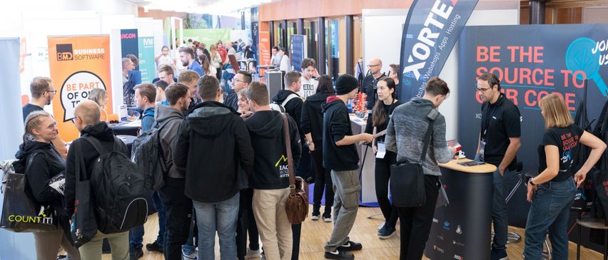 Eine Menschenmenge steht in einer Schlange, wahrscheinlich auf einer Konferenz. Einige tragen Rucksäcke und Brillen. Sie stehen vor einem Stand mit einem blauen Banner. Der Boden ist aus Holz und die Wände sind aus Holz und Glas.