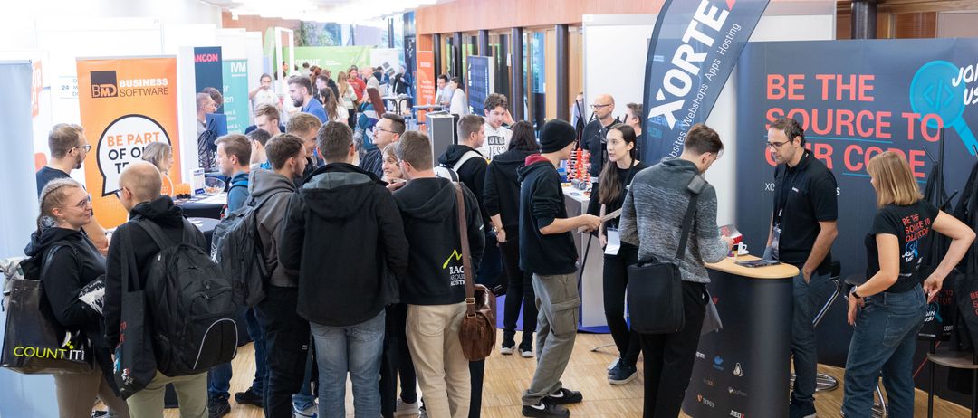 Eine Menschenmenge steht in einer Schlange, wahrscheinlich auf einer Konferenz. Einige tragen Rucksäcke und Brillen. Sie stehen vor einem Stand mit einem blauen Banner. Der Boden ist aus Holz und die Wände sind aus Holz und Glas.