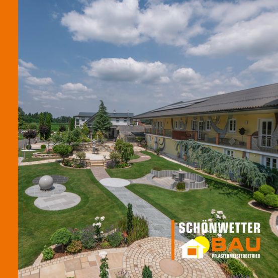 Luftaufnahme eines gepflegten Gartens mit einem Gebäude mit gelber Fassade und grauem Dach. Der Garten umfasst verschiedene Pflanzen, Bäume und einen Betonweg. Ein Wasserzeichen mit dem Text 'SCHONWETTERBAU' befindet sich in der unteren rechten Ecke.