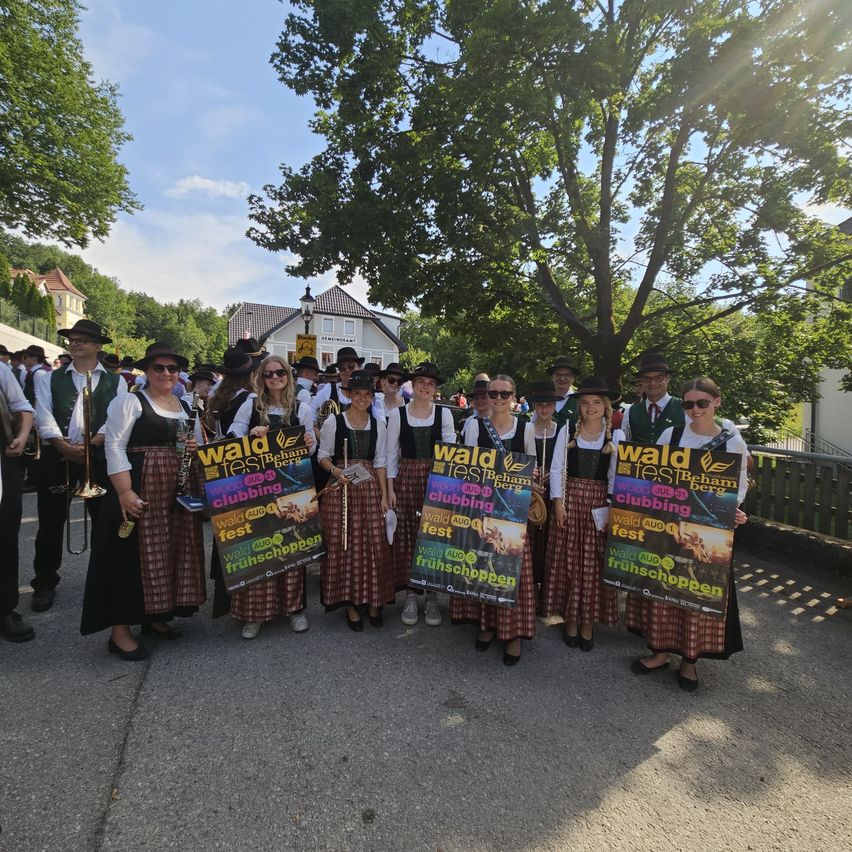 Eine Gruppe von Menschen in traditioneller Kleidung posiert für ein Foto mit Bannern. Sie stehen an einem sonnigen Ort im Freien, möglicherweise bei einem Festival.