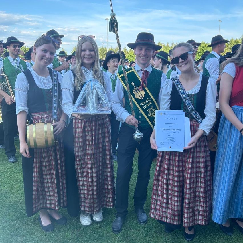 Gruppe von Menschen in traditioneller Kleidung bei einem Festival. Zwei Frauen halten ein Zertifikat und eine Trophäe, während andere Musikinstrumente spielen.
