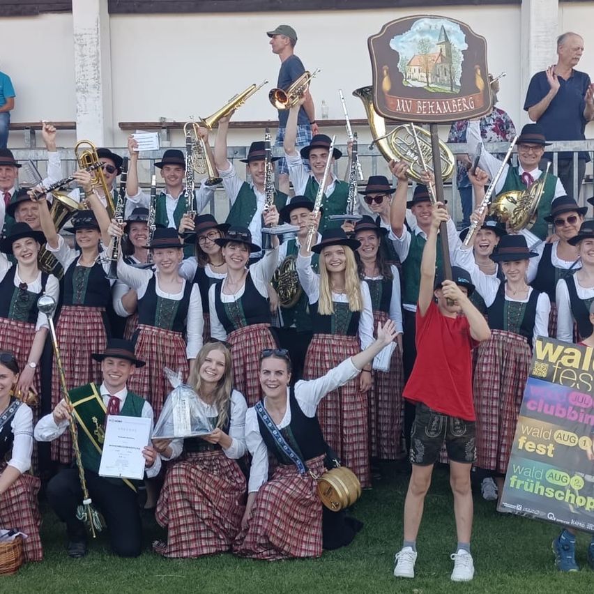 Eine Gruppe von Menschen in traditionellen Kostümen hält Musikinstrumente und Trophäen. Sie lächeln und posieren für ein Foto bei einer Outdoor-Veranstaltung.