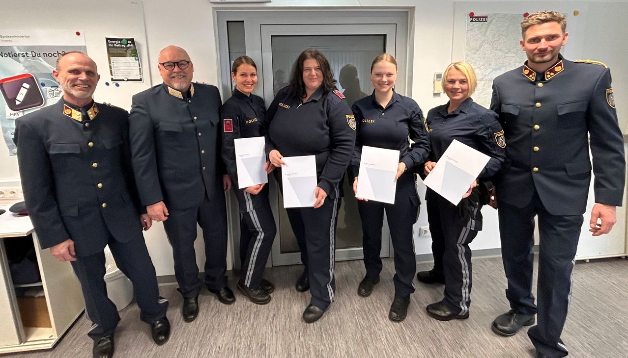Fünf Personen in Polizeiuniformen stehen nebeneinander und halten Urkunden in der Hand, möglicherweise für ein Foto in einem Büroraum posierend.