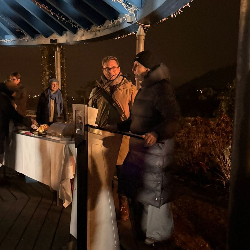 Zwei Personen stehen bei Nacht auf einer Terrasse am Rednerpult mit Mikrofonen. Eine Person hält ein Mikrofonstativ, während die andere spricht. In der Nähe steht ein Tisch mit Essen.