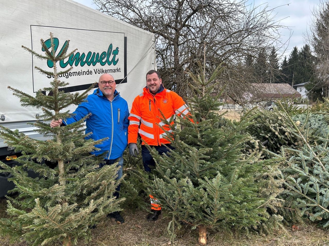 Zwei Männer stehen zwischen Kiefern, einer trägt einen blauen Mantel, der andere einen orangefarbenen reflektierenden Mantel. Sie sind draußen auf einem Feld. Hinter ihnen steht ein weißer LKW mit einem Schild, auf dem 'Unnwelt' steht.