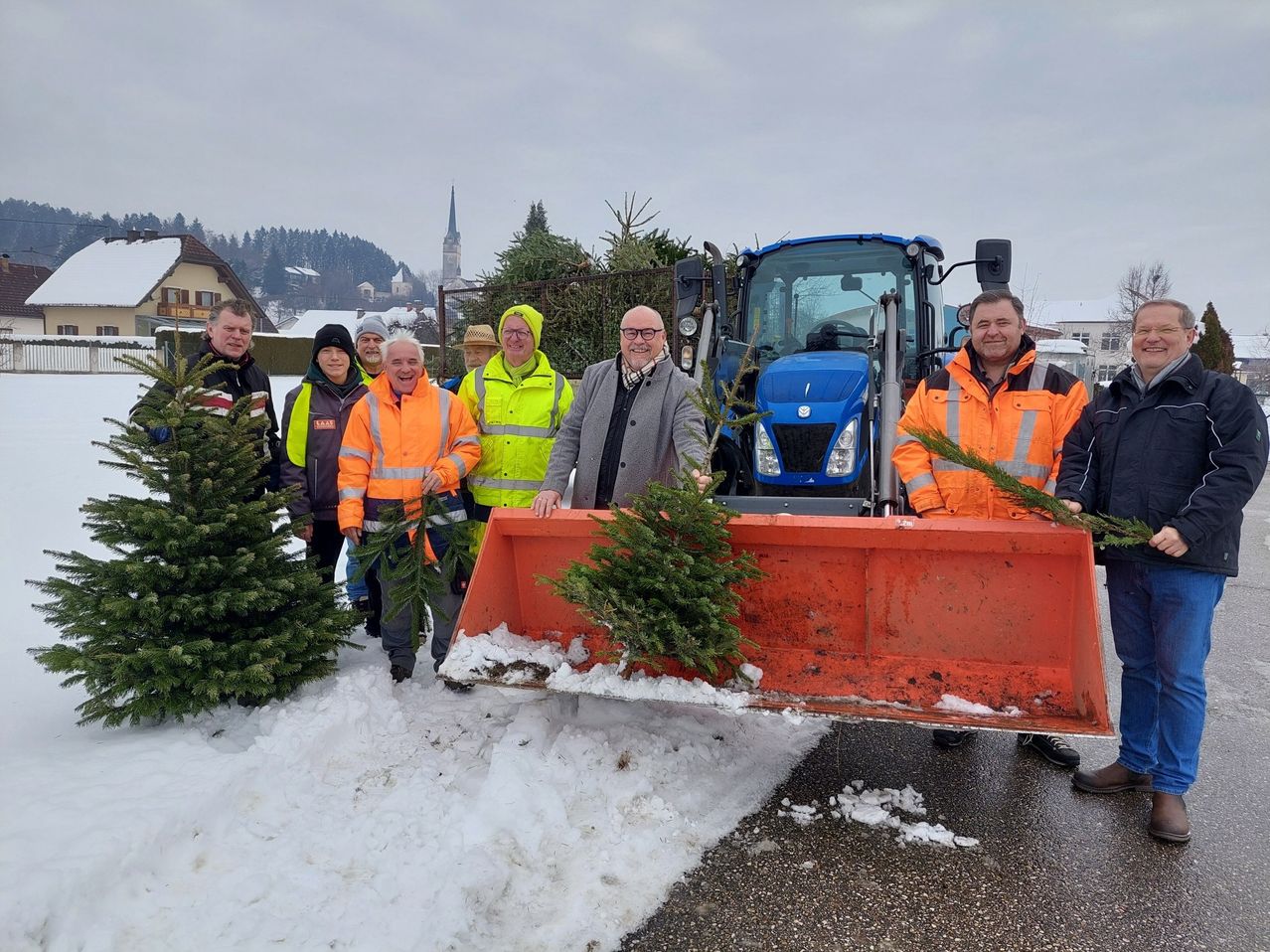 Eine Gruppe von Männern in Warnwesten steht neben einem Traktor mit Weihnachtsbäumen im Schnee. Sie halten Tannenzweige.