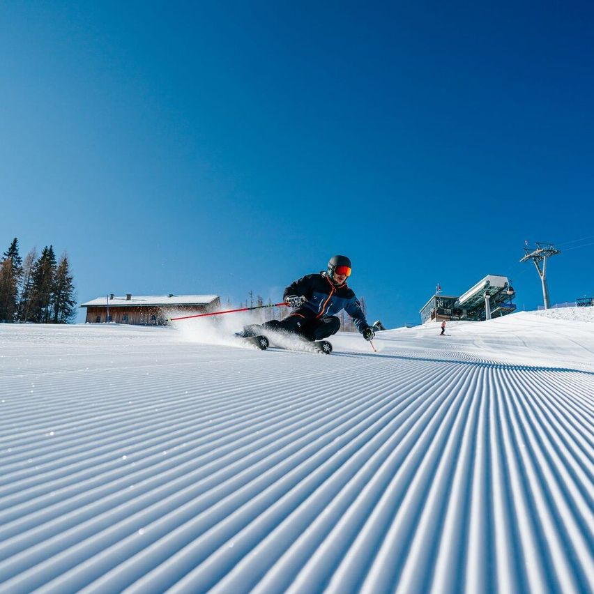 Ein Skifahrer fährt einen schneebedeckten Hang unter einem klaren blauen Himmel hinunter. Bäume und eine Skilift sind im Hintergrund zu sehen.