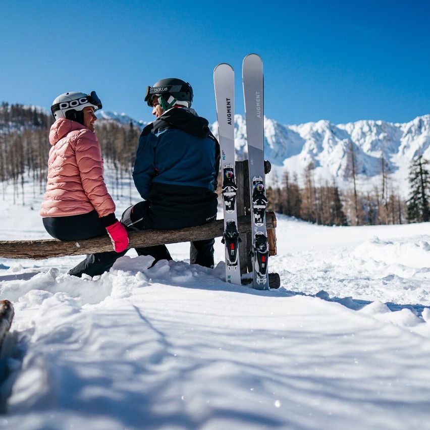 Zwei Skifahrer, eine Frau und ein Mann, sitzen auf einem Baumstamm im Schnee und tragen Skiausrüstung. Hinter ihnen stehen Skier mit der Marke Augment. Im Hintergrund sind verschneite Berge zu sehen.