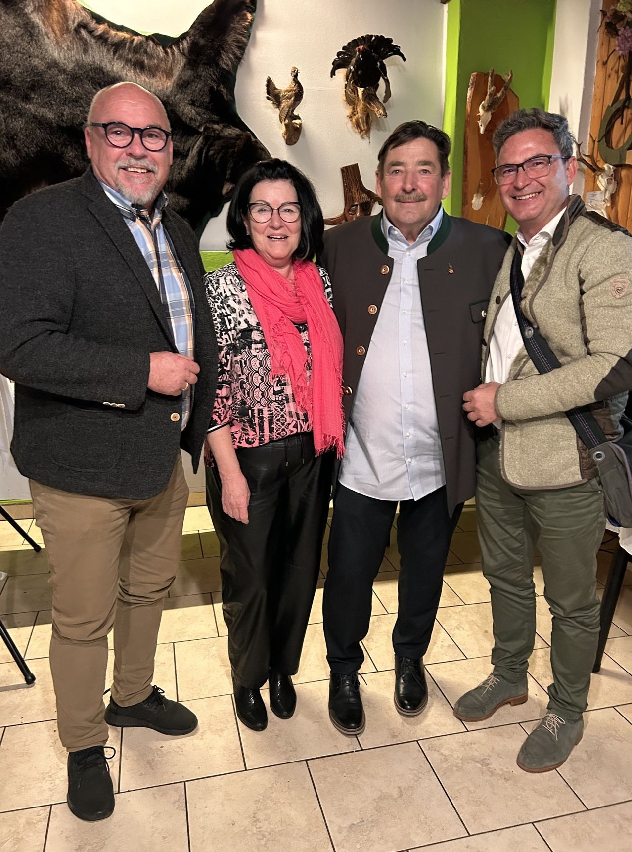Four adults, two men and two women, are standing together indoors. They are smiling and seem to be posing for a photo. The woman in the middle is wearing a scarf. Behind them, there is a wall with a bear head and some animal trophies.