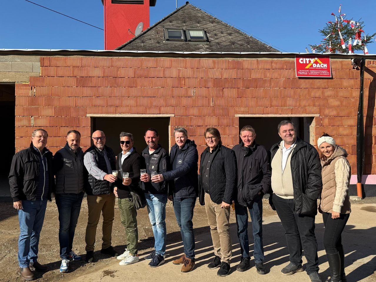 A group of men and a woman stand in front of a red building with a sign reading City Dach. They are all dressed warmly and smiling, with some holding glasses. Behind them, a tree is visible in the distance.