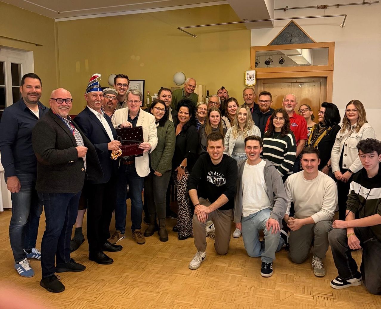 A group of people are posing for a picture, some standing and others sitting, in a room with a wooden floor and yellow walls. A man is holding a trophy, and a few people are wearing glasses.