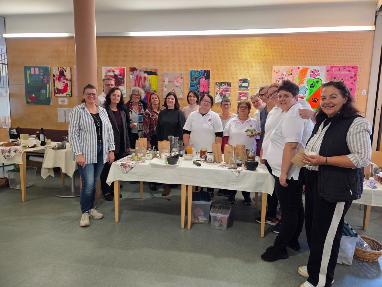 A group of people stand around a table with various items, smiling. Some wear white shirts, others casual clothing. Behind them, artworks hang on the wall.