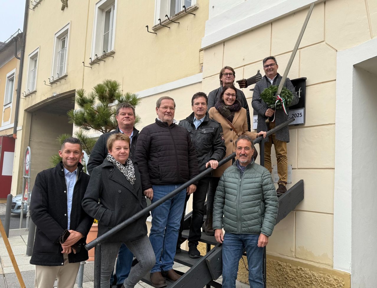 A group of people pose for a picture on a building's staircase, with one holding a wreath and sword. The building has white windows and a sign reading '30.2025'.