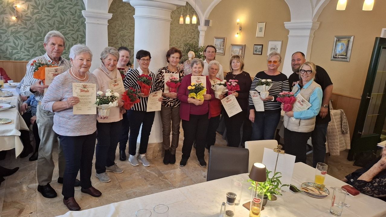 A group of women and men are posing for a photo with flowers and certificates in their hands. They are smiling and standing in a room.