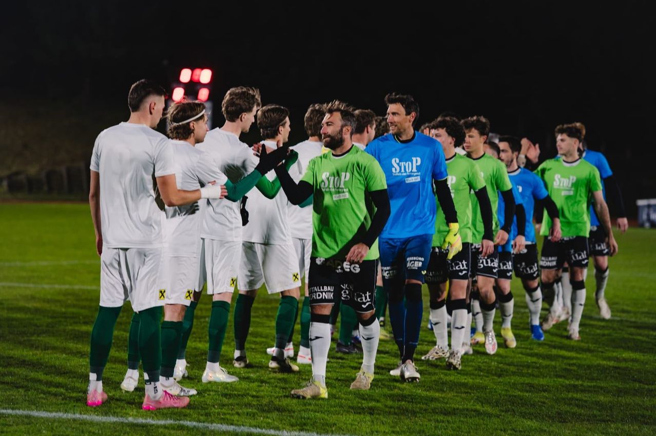 Fußballspieler in grünen und blauen Trikots laufen bei Nacht vom Feld und lächeln. Sie schütteln Hände. Das Feld hat eine Anzeigetafel mit roten Lichtern.