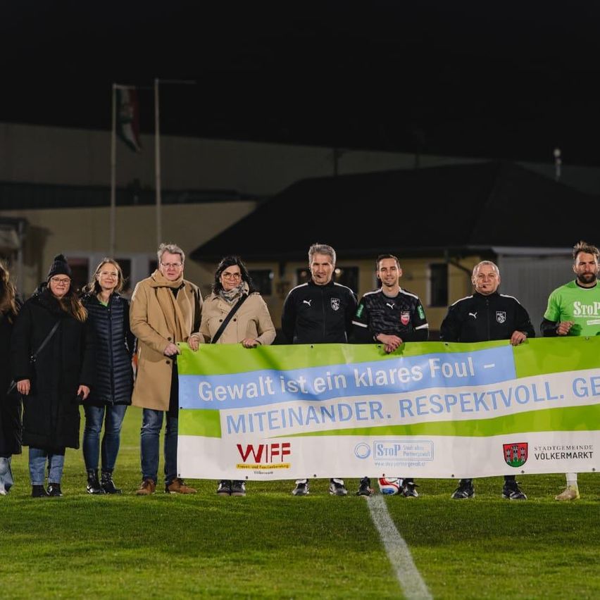 Eine Gruppe von Erwachsenen und Kindern steht auf einem Fußballfeld und hält ein Banner. Das Banner trägt die Aufschrift 'Gewalt ist ein klares Foul - Miteinander. Respektvoll. Gewissenhaft.' Es gibt mehrere Personen in schwarzen Trikots mit Nummern.