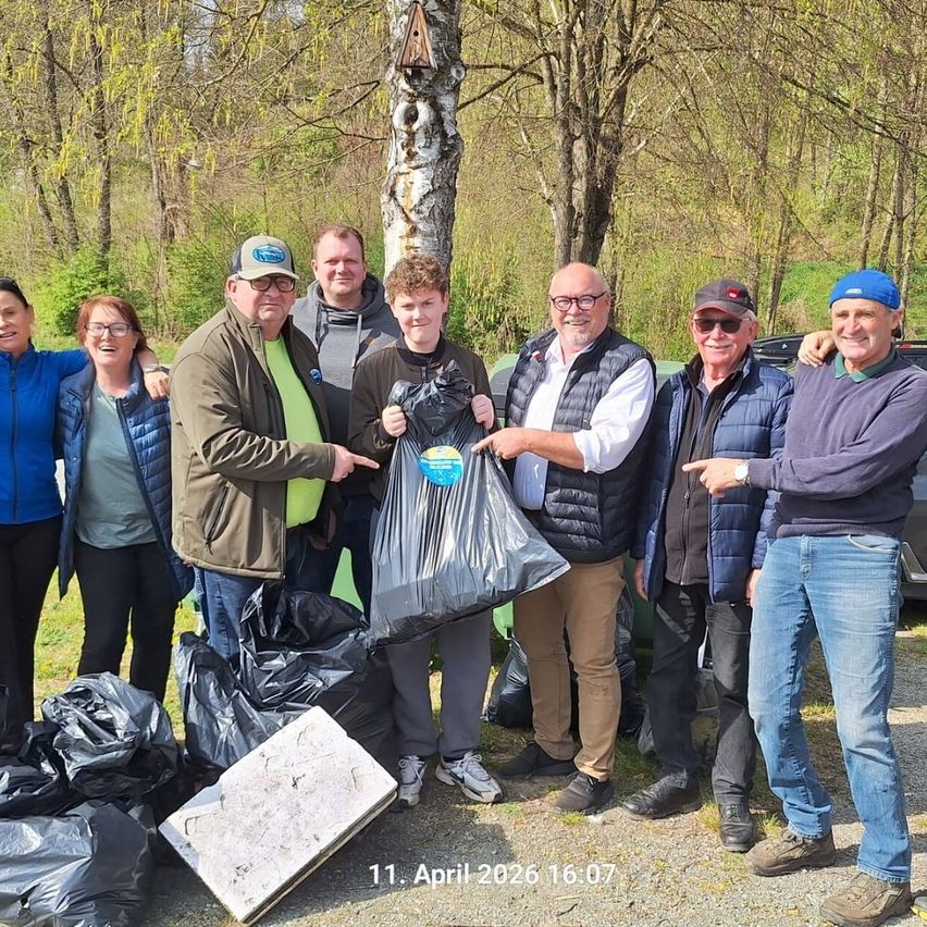 Eine Gruppe von Menschen versammelt sich im Freien, einige mit Taschen, posieren für ein Foto in einem bewaldeten Gebiet am 11. April 2026.