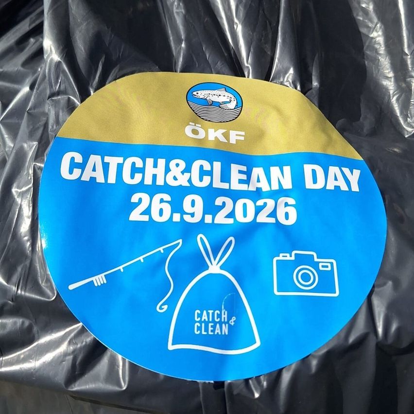 Rundes blaues Schild mit 'OeKF CATCH & CLEAN DAY 26.9.2026' darauf geschrieben. Es hat ein Fischlogo, eine Angel, einen Müllsack und ein Kamerasymbol. Das Schild ist auf Plastik.