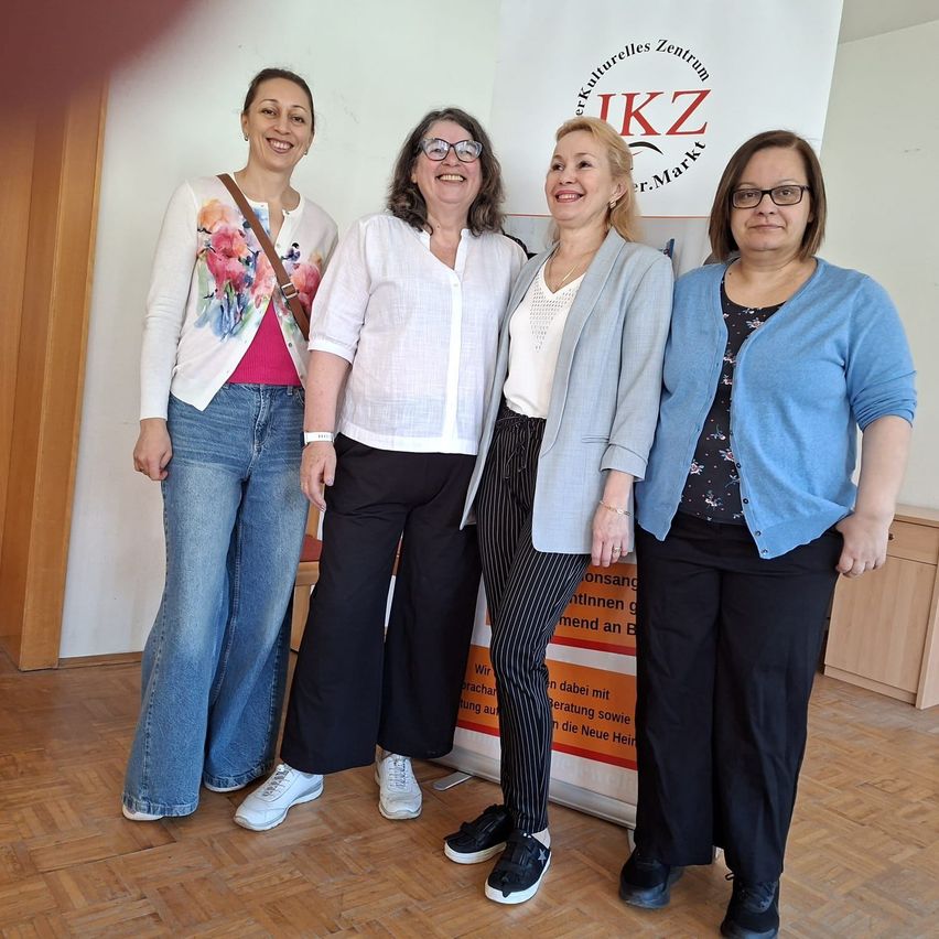 Vier Frauen stehen lächelnd zusammen in einem Raum, einer hält eine Tasche. Sie stehen vor einem Banner. Sie tragen Freizeitkleidung.