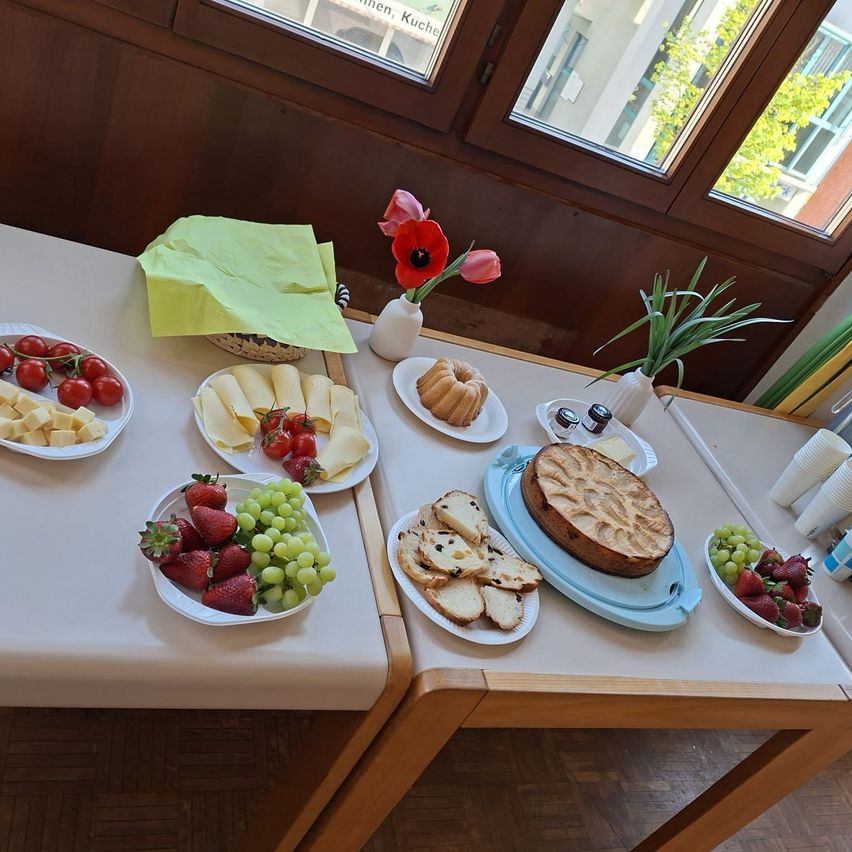 Ein Tisch zeigt eine Auswahl an Speisen, darunter Käse, Tomaten, Erdbeeren, Trauben und Brot auf zwei Tellern. Es gibt Blumen in einer Vase und Papierservietten auf dem Tisch.