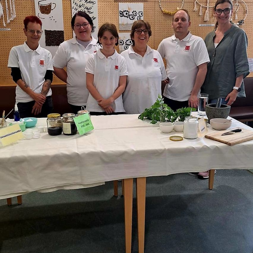 Sechs Menschen posieren für ein Foto bei einer Veranstaltung. Fünf tragen weiße Hemden mit Logos und stehen hinter einem Tisch mit verschiedenen Gegenständen. Eine Frau ist in einem grauen Hemd und lächelt.