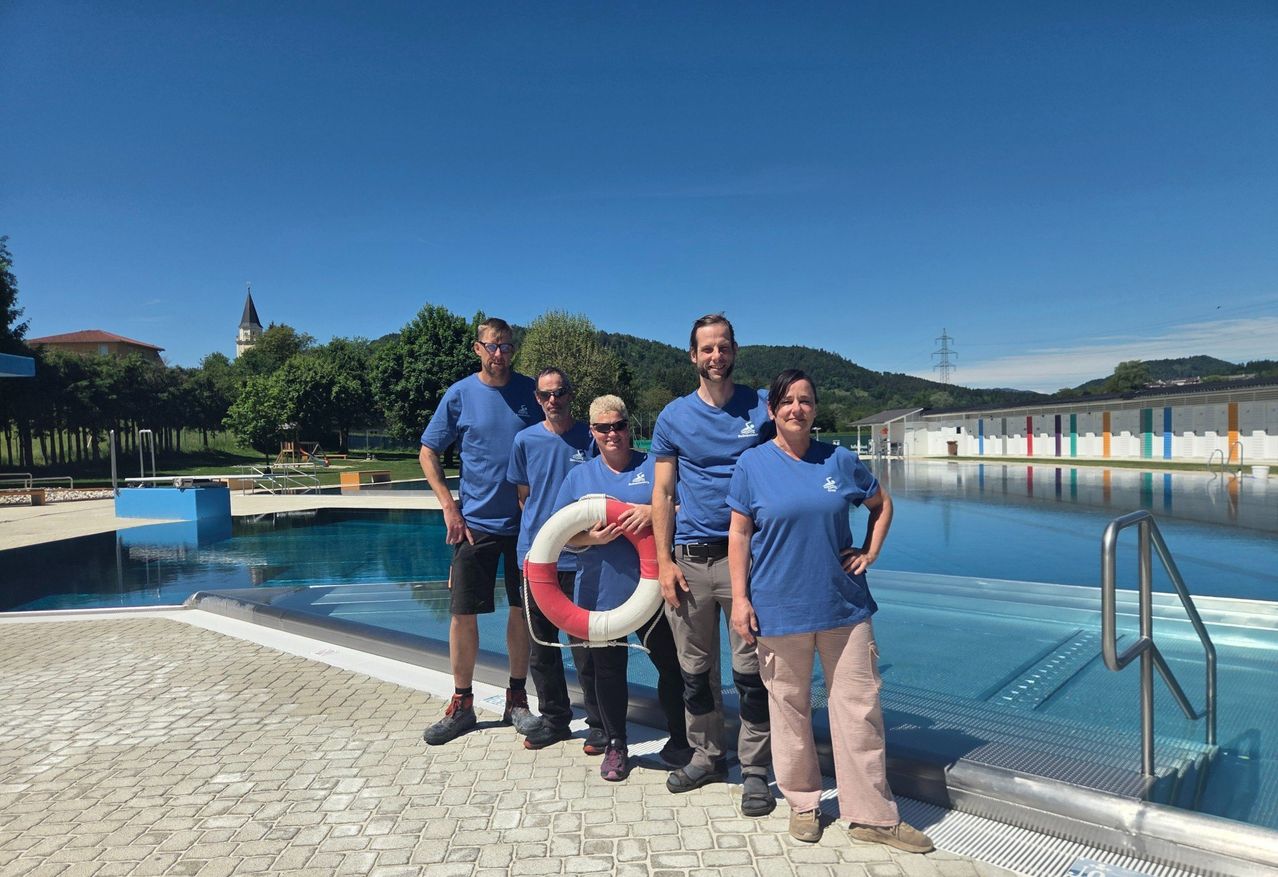 Fünf Personen stehen neben einem Schwimmbecken, alle in blauen T-Shirts gekleidet. Eine Person hält einen Rettungsring. Hinter ihnen steht ein Gebäude und es gibt Bäume. Der Himmel ist klar.