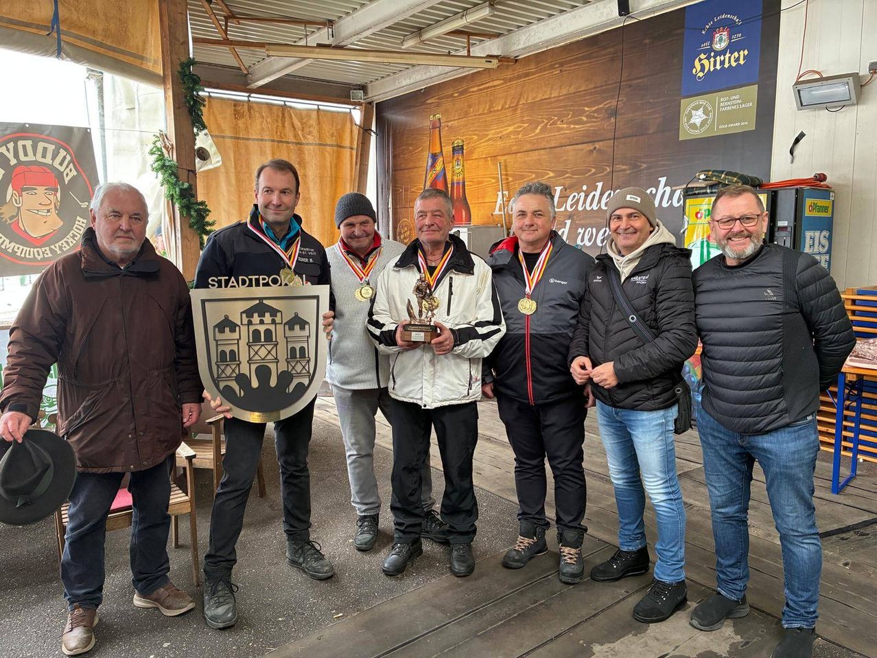 Mehrere Männer stehen in einem Innenbereich zusammen, einige mit Medaillen. Ein Mann hält eine Trophäe und einen Schild. Sie alle lächeln.